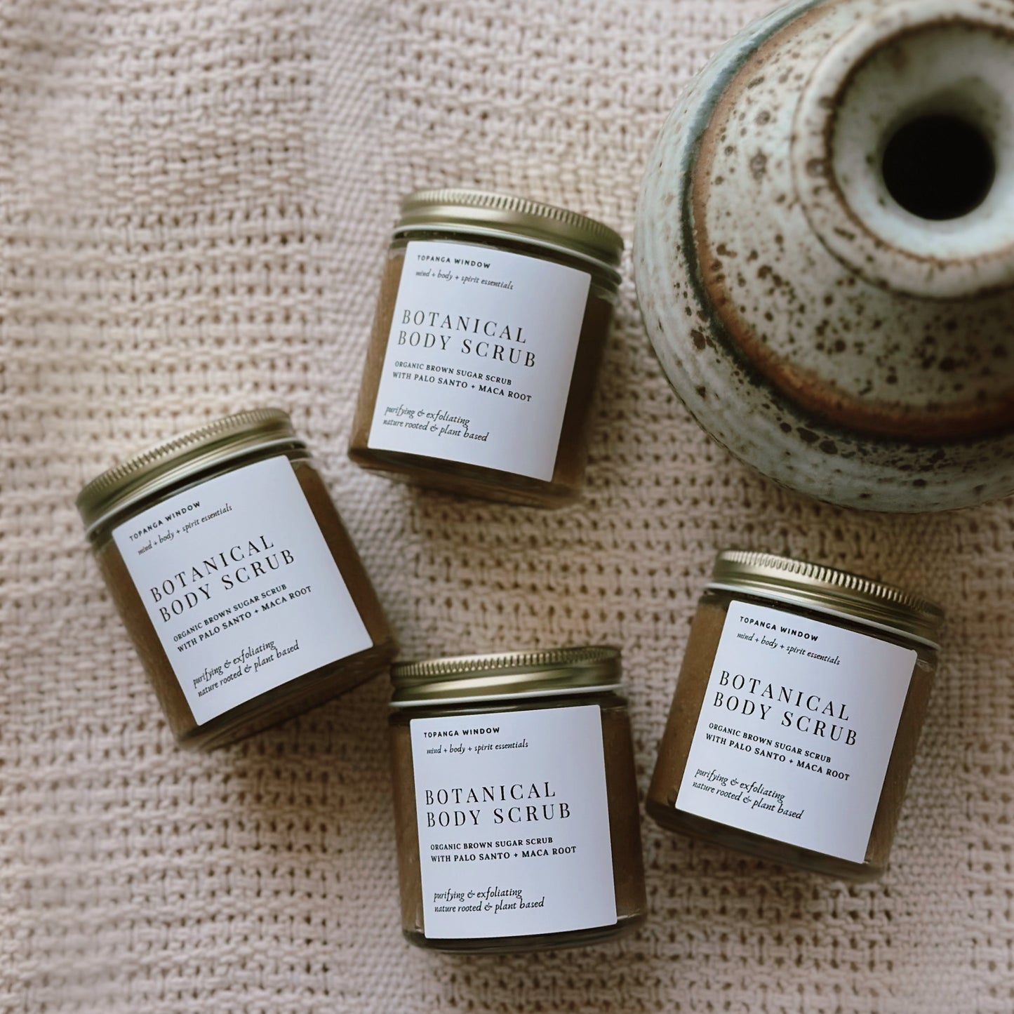 Four jars of Botanical Body Scrub on a textured surface with a ceramic container.