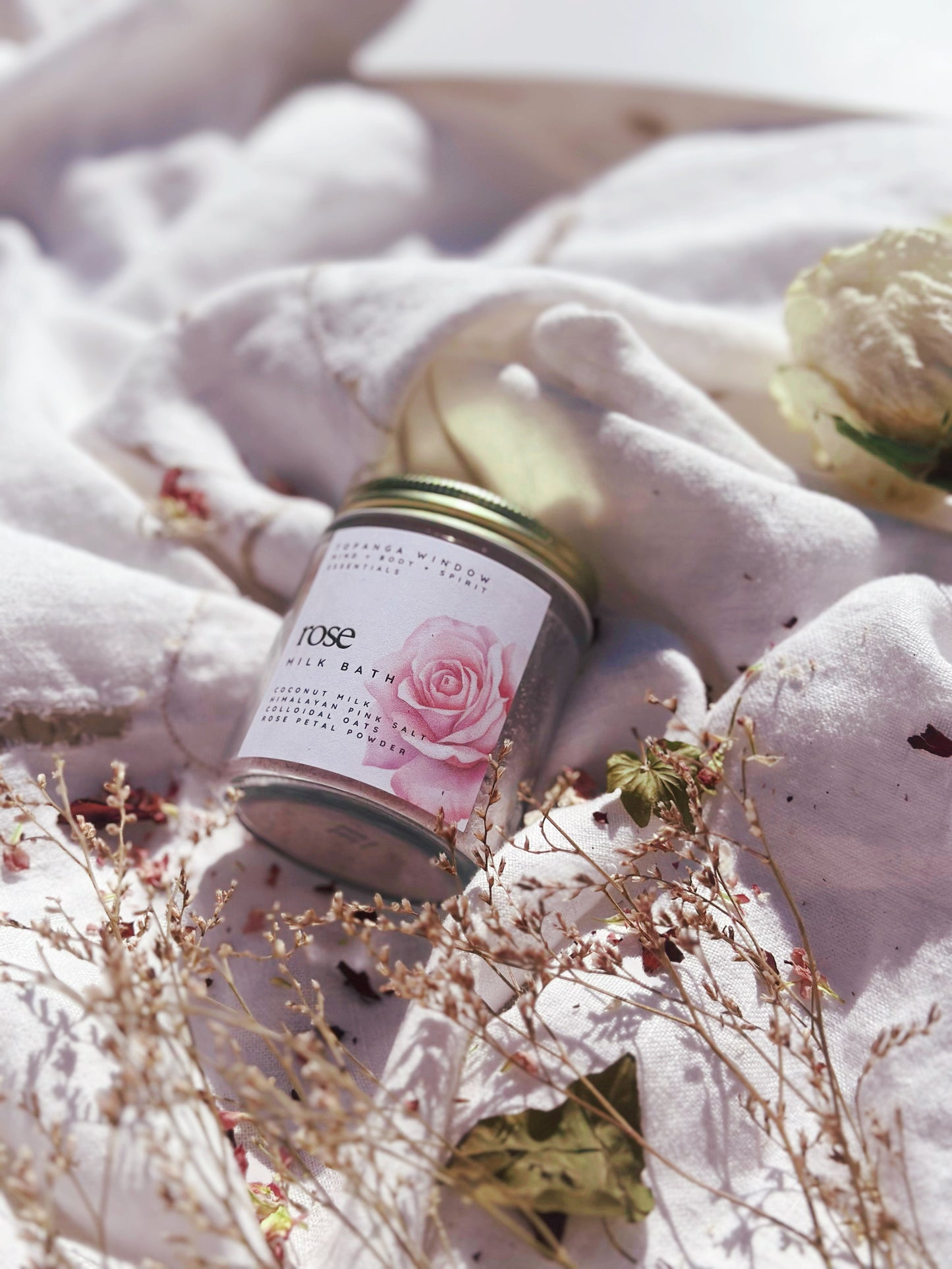 glass jar of rose milk bath soak on soft white cloth with scattered dried florals