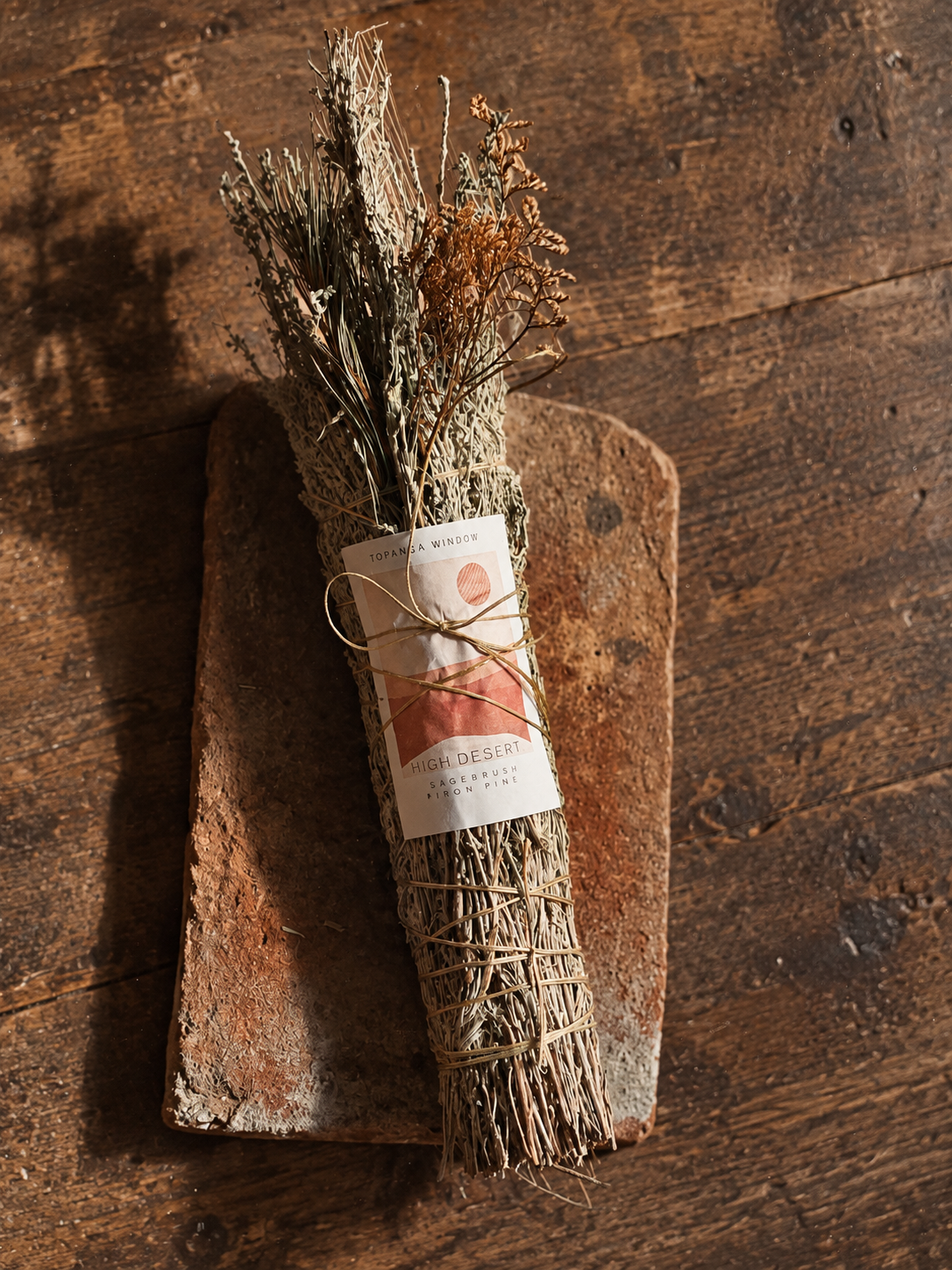 A dried botanical sagebrush bundle wrapped in twine with a paper label reading High Desert and '100% ORGANIC', resting on a rustic wooden board over a dark wood tabletop.