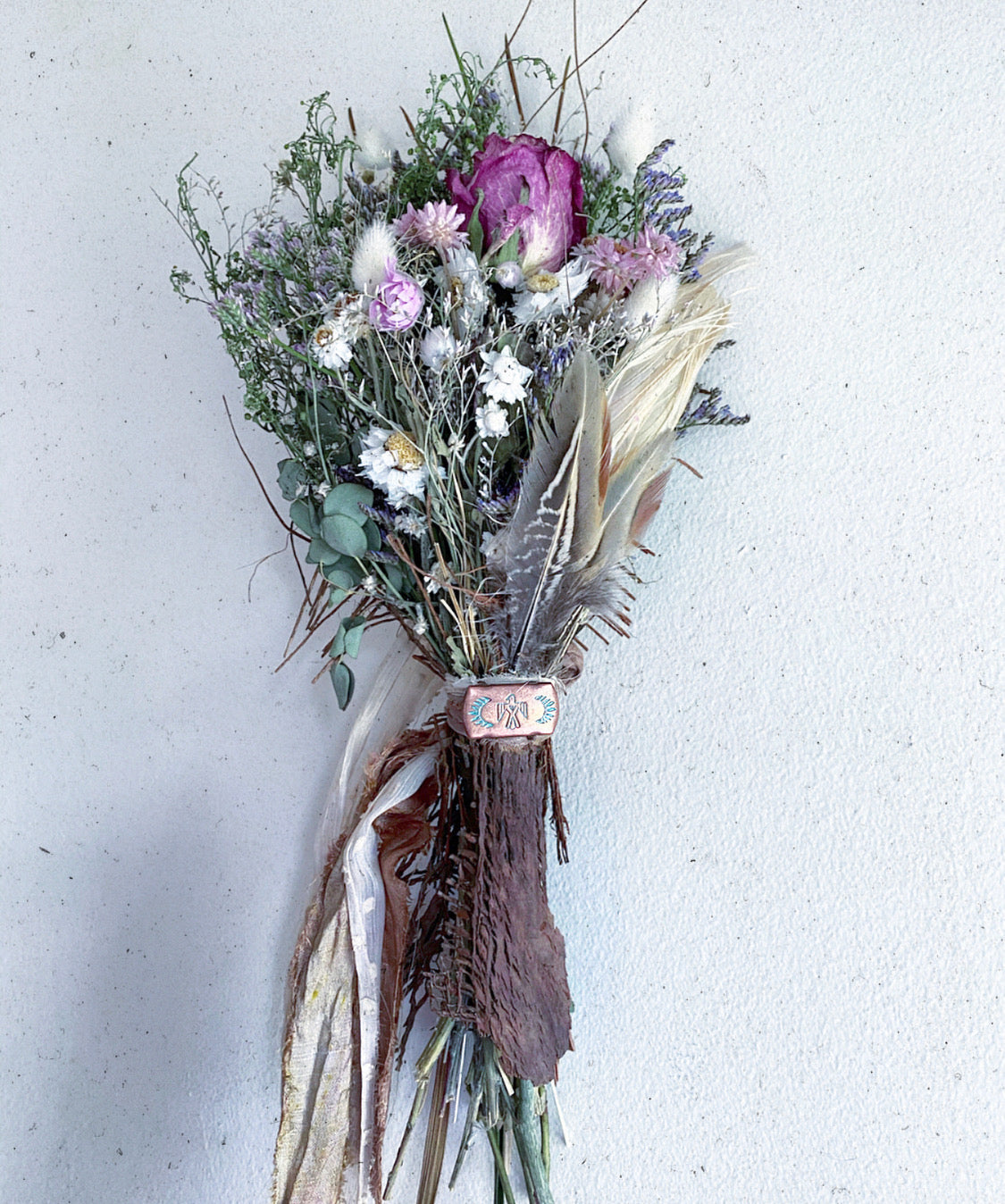 A dried flower bouquet featuring eucalyptus, chamomile, sea lavender, purple wild rose, bunny tails, amaranth, and wildflowers with added pheasant feathers and a Thunderbird concho.