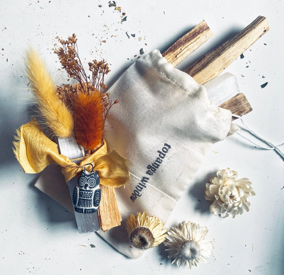 A set of Palo Santo sticks, white selenite crystals, and a Bolivian good luck amulet, all arranged on a surface with a yellow silk ribbon and a storage drawstring bag.