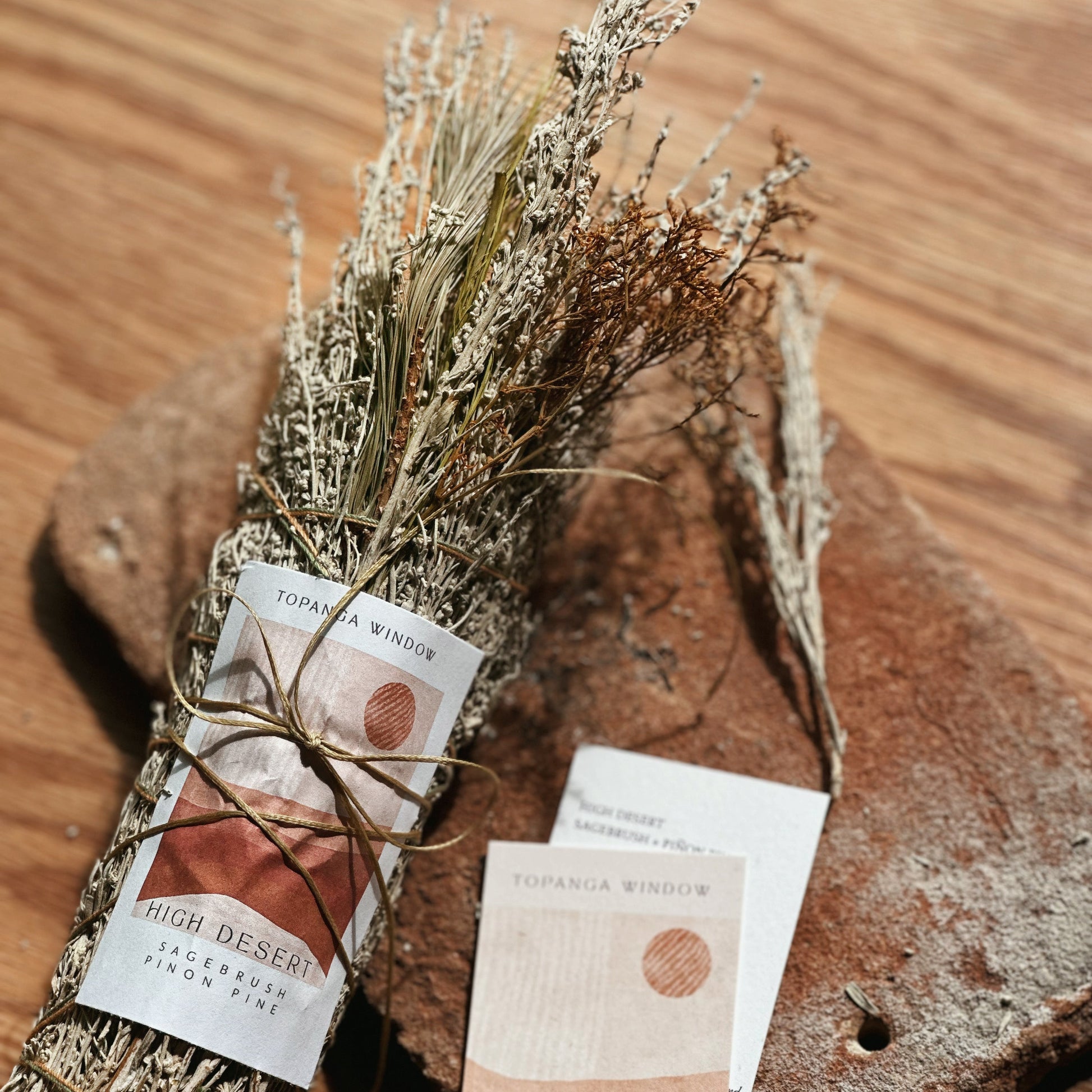 A bundle of dried botanicals tied with twine and a matching printed card, placed on a rustic wooden surface with a textured stone prop. The card reads 'HIGH DESERT' and TOPANGA WINDOW