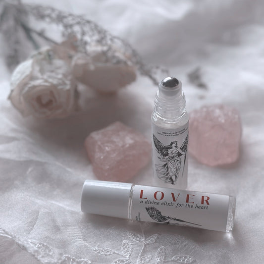 A glass roll-on bottle of 'LOVER' perfume oil, placed on a textured white surface, with decorative elements including a rose and crystals in the background.