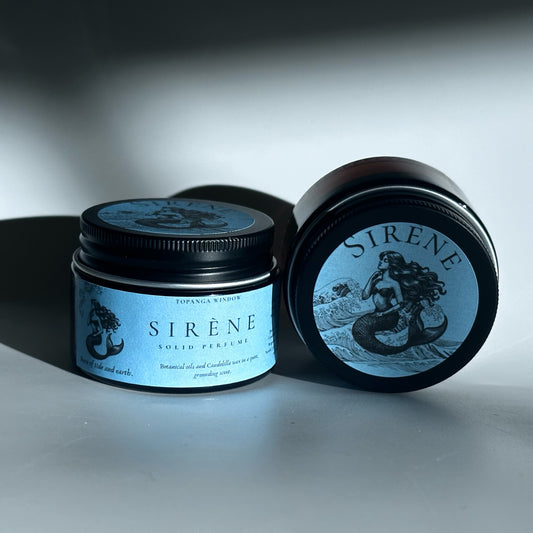 Two 'SIRENE' solid perfume containers on a reflective surface with a dark background