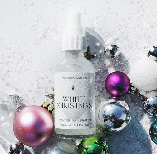 A clear glass bottle labeled 'White Christmas Holiday Room Mist' by Topanga Window, surrounded by holiday decorations including Christmas balls and ornaments on a white background.