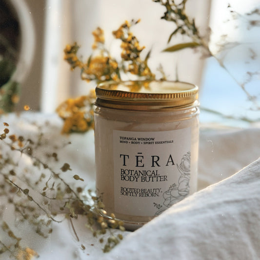 Téra Botanical Body Butter jar with gold lid on a white surface with dried flowers.