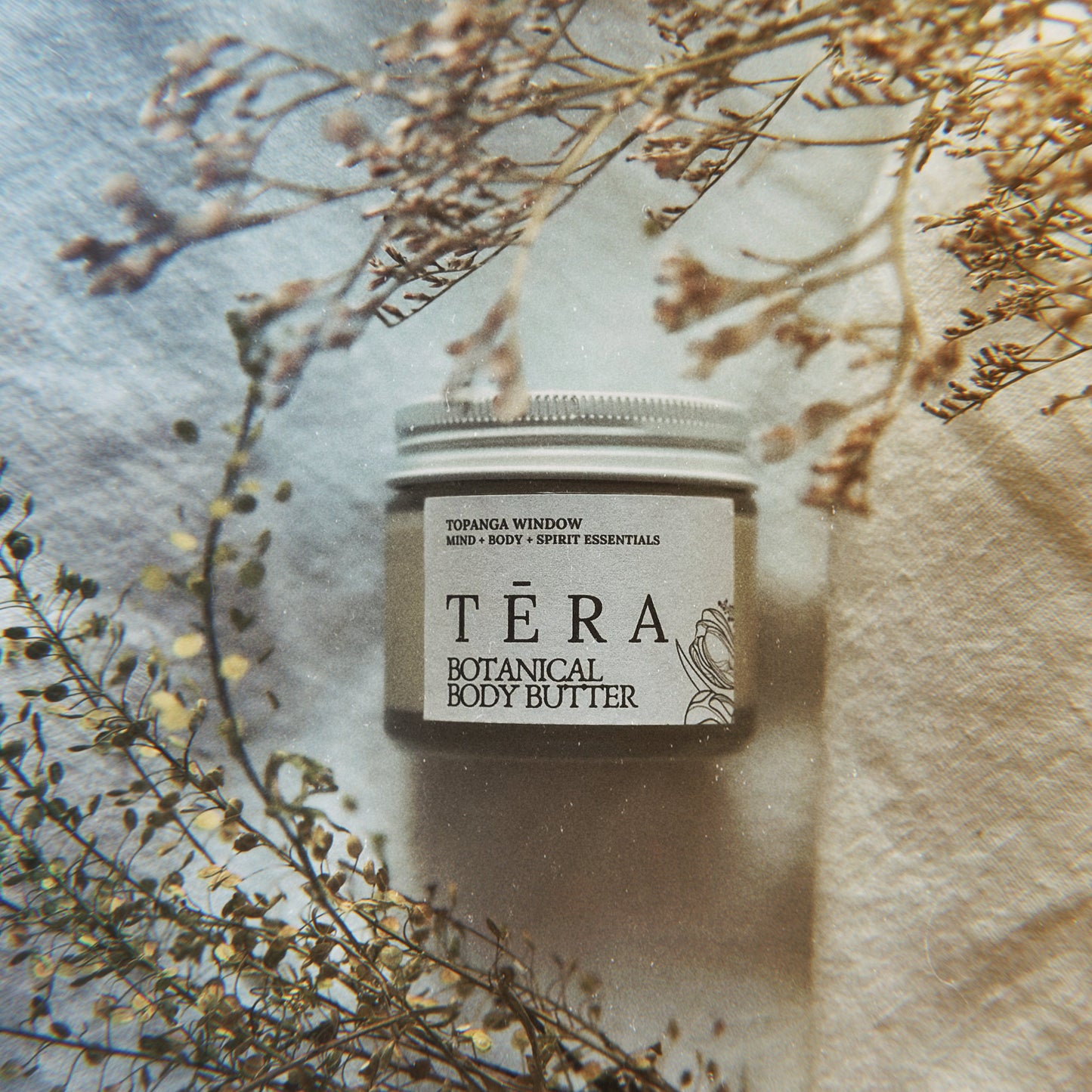 Téra Botanical Body Butter jar with dried flowers on a textured fabric background