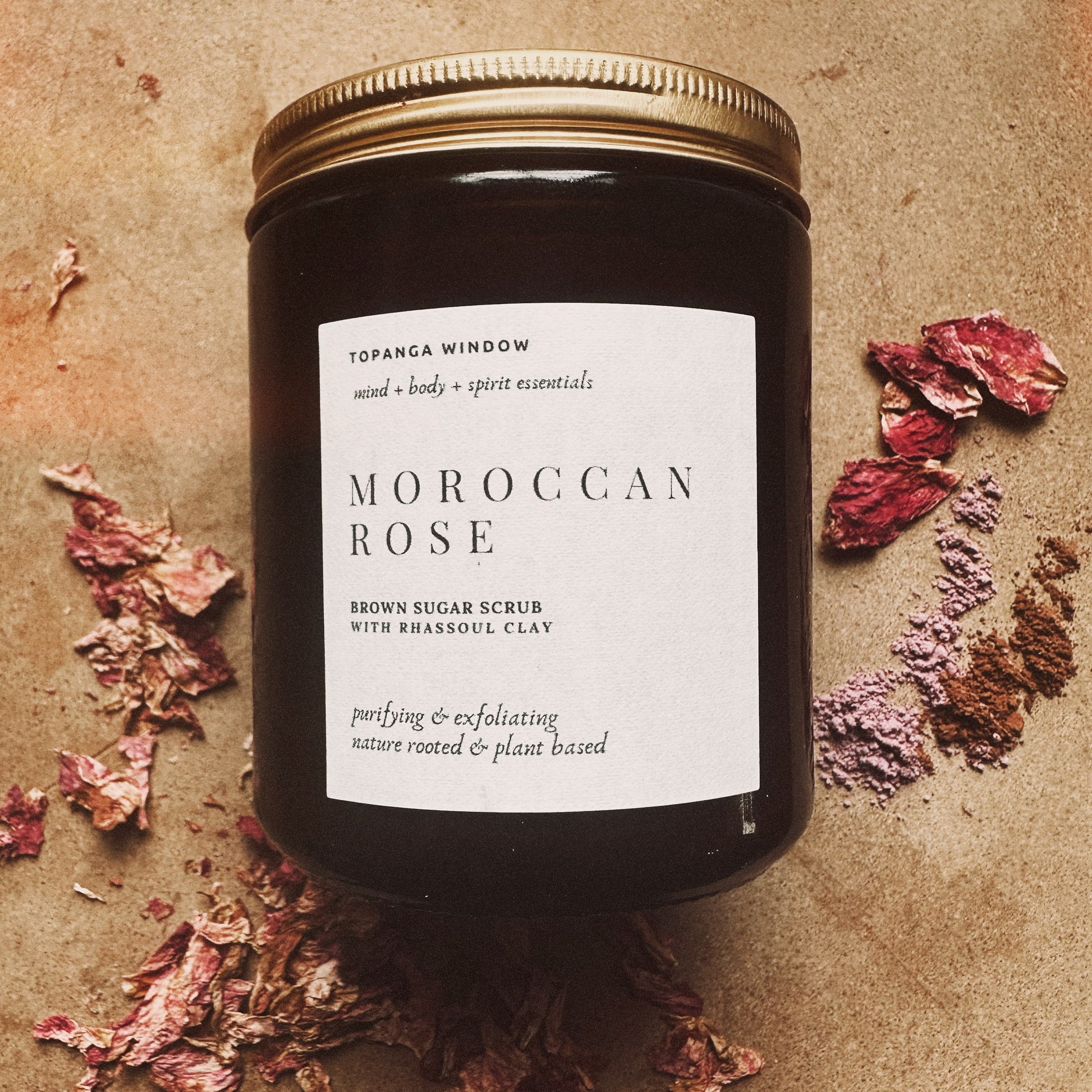 Jar of Moroccan Rose brown sugar scrub with rhassoul clay on a textured surface with dried flowers.