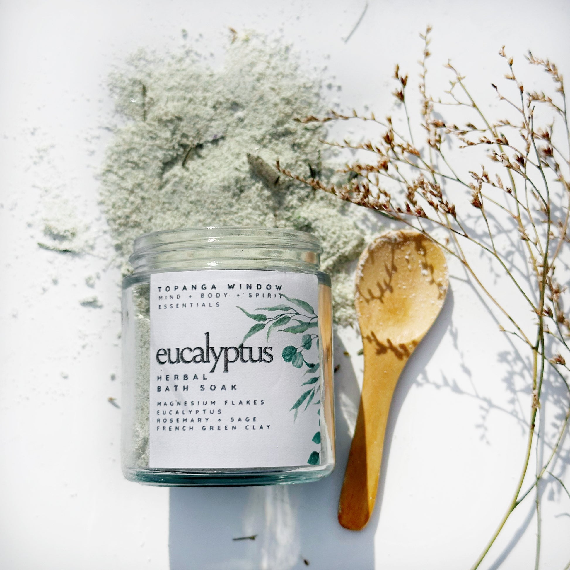 A jar of EUCALYPTUS HERBAL BATH SOAK with a wooden spoon and a muslin bag placed on top, against a white background.