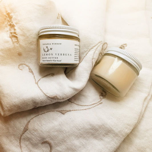 Two small jars of body butter on a textured fabric background