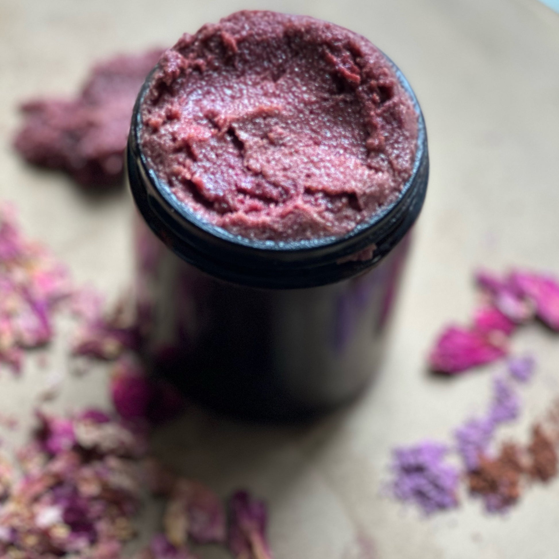 Black container with purple sugar scrub on a surface with dried flowers