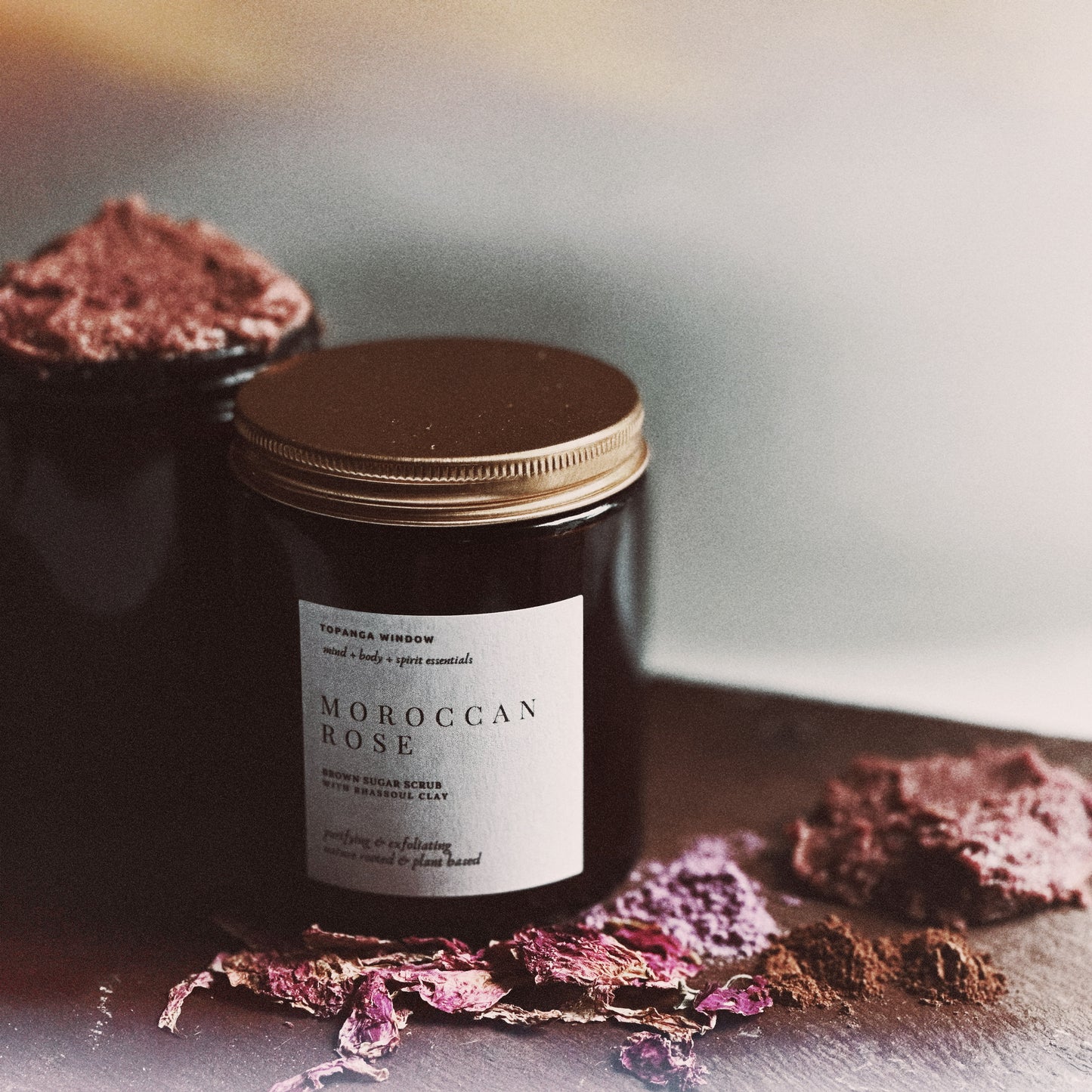 Jar of Moroccan Rose scrub with a gold lid on a reflective surface with dried flowers.