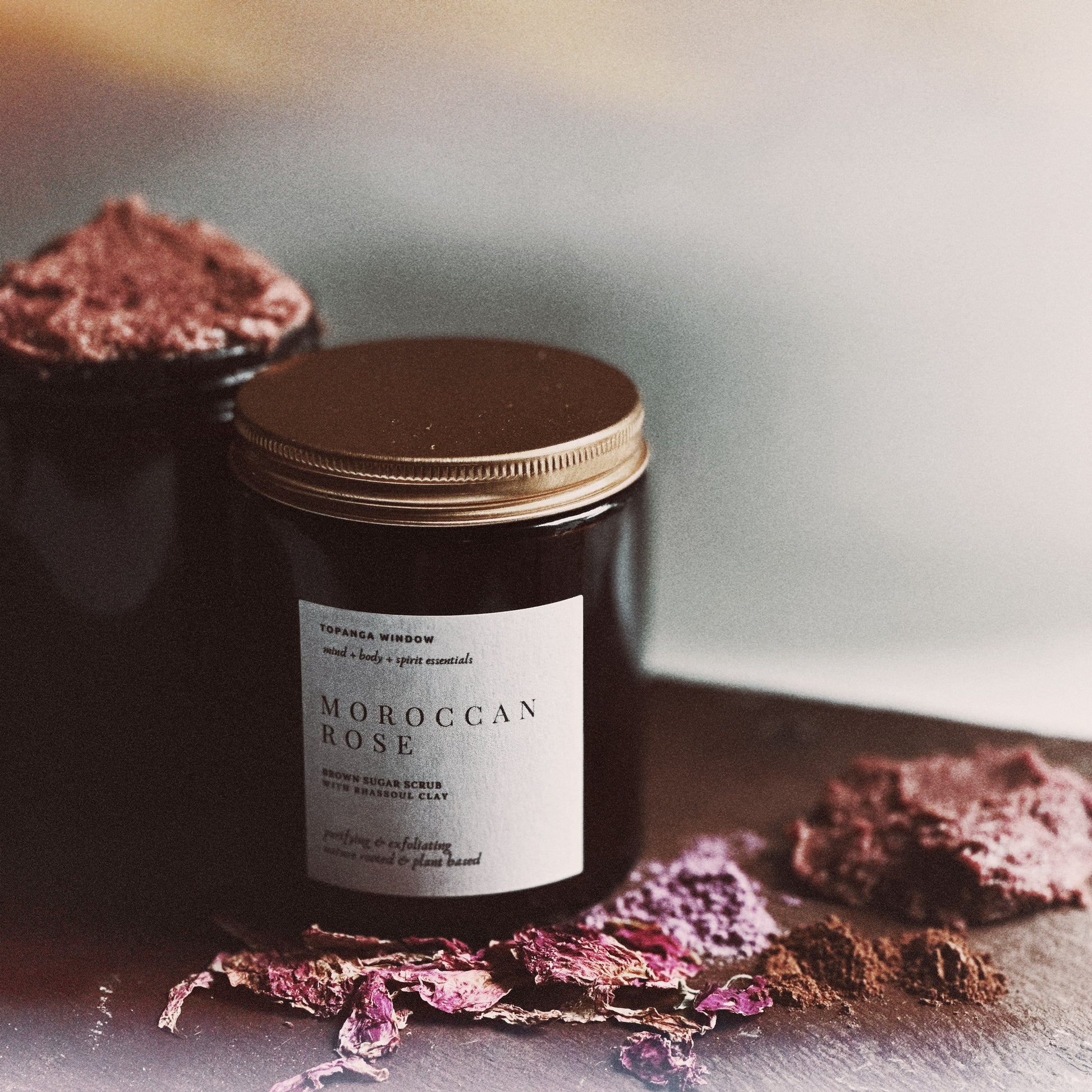 Jar of Moroccan Rose scrub with a gold lid on a reflective surface with dried flowers.