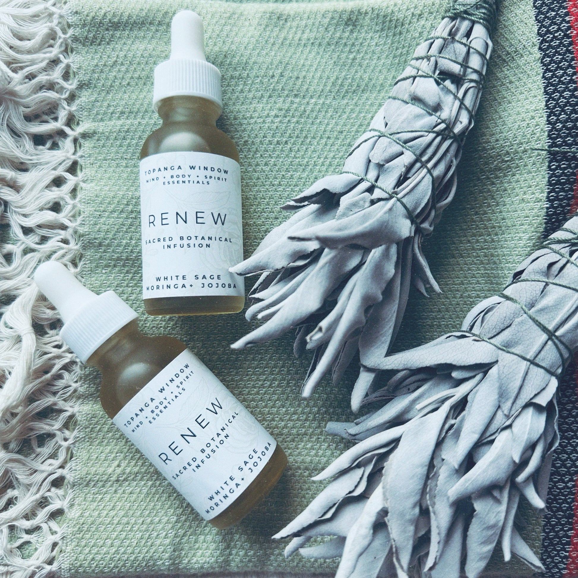 Two glass dropper bottles of RENEW White Sage Sacred Oil on a textured fabric surface, alongside a bundle of white sage.