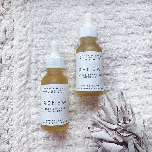 Two small glass dropper bottles labeled 'RENEW White Sage Sacred Oil', with white caps and branding on a textured white background with a dried white flower beside them.
