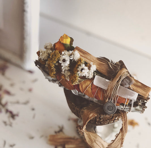 yerba santa stick used for spiritual smoke cleansing . Bundled together with dried orange rose petals, yarrow, white flowers and yellow roses.  with gold and orange silk ribbons sitting on a gold dish on a white table.