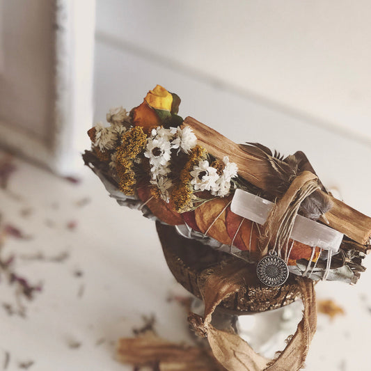 yerba santa stick used for spiritual smoke cleansing . Bundled together with dried orange rose petals, yarrow, white flowers and yellow roses. with gold and orange silk ribbons sitting on a gold dish on a white table.